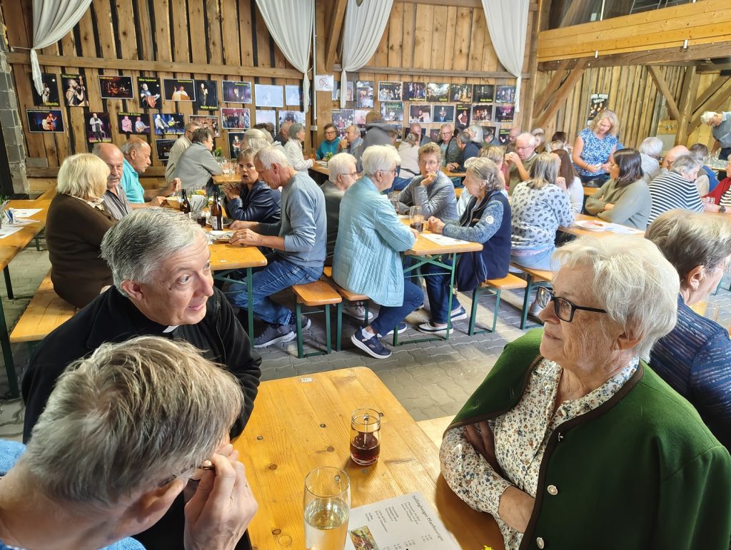 Menschen sitzen an Holztischen in einem Stall, im Hintergrund Bilder von Musikern.